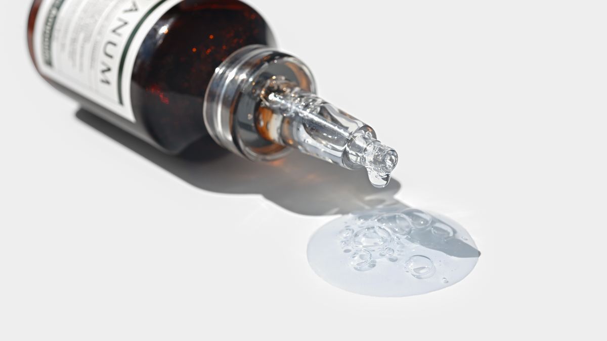 Korean ampoule bottle lying on a white surface, liquid pooling from its tapered glass tip